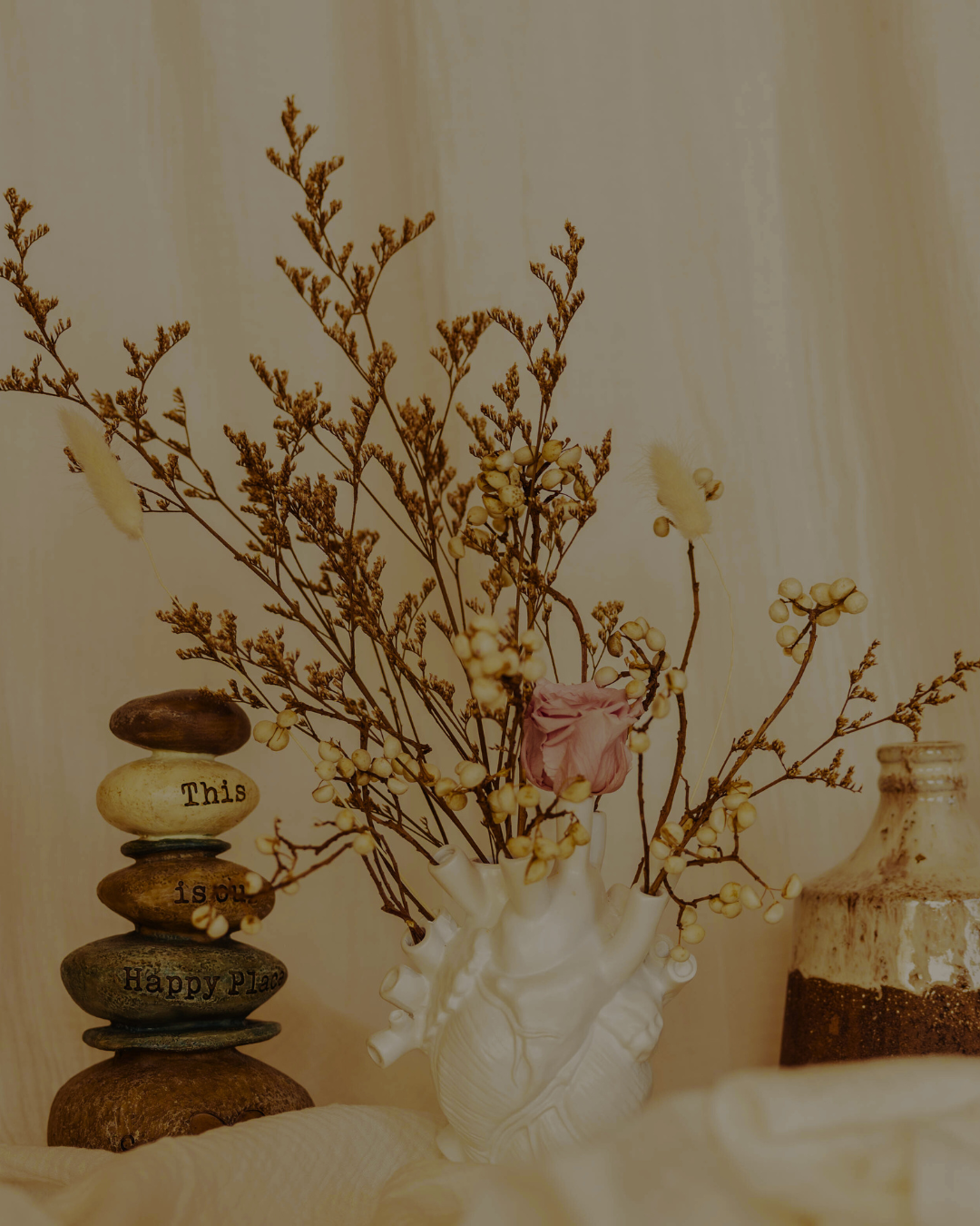 a dried flowers including dried cotton branch, and pink dried rose in white heart shaped vase with stacking rocks and a clay vase in the back