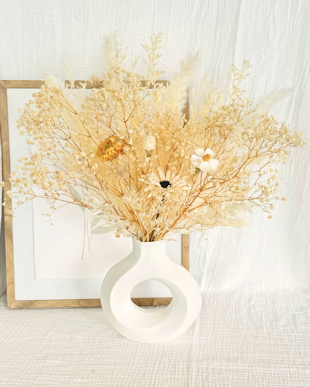 a white twisted donut shaped vase with white themed dried flower display on a table